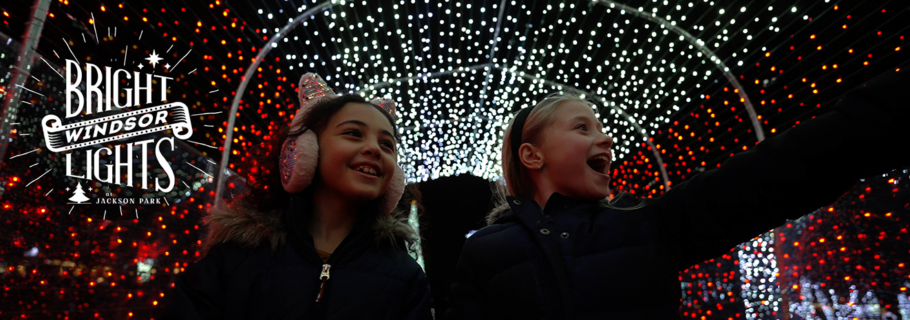 Logo for Bright Lights Windsor at Jackson Park and two smiling young girls in a light tunnel, one with sparkly cat earmuffs, one pointing at the lights in awe